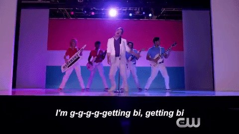 A gif of Darryl Whitefeather dressed in a white tuxedo and dancing in front of the bi pride flag with the lyrics, "I'm g-g-g-g-getting bi. I'm getting bi," below him.